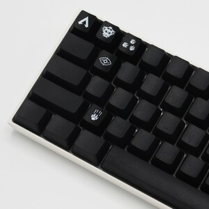 Peut inclure: Un clavier m&eacute;canique noir avec des touches blanches comportant divers symboles, dont un cr&acirc;ne, un &oelig;il et des empreintes de pattes. Le clavier a un cadre blanc et une barre d'espace noire.