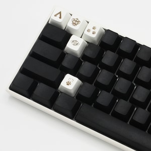 Peut inclure: Un clavier m&eacute;canique noir avec des touches blanches comportant divers symboles, notamment un triangle, un &oelig;il et des empreintes de pattes. Le clavier a une barre d'espace noire et un cadre blanc.