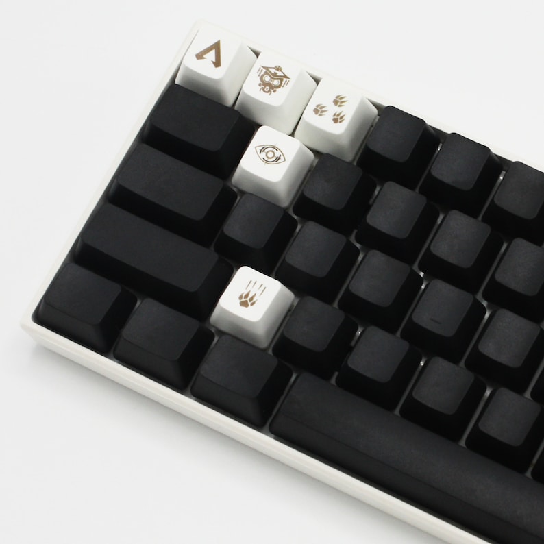 Peut inclure: Un clavier m&eacute;canique noir avec des touches blanches comportant divers symboles, notamment un triangle, un &oelig;il et des empreintes de pattes. Le clavier a une barre d'espace noire et un cadre blanc.