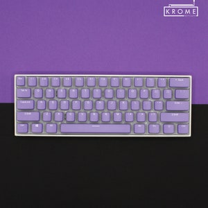 May include: A compact, white-framed keyboard with lavender keycaps. The keys are arranged in a standard layout, with letters, numbers, and symbols. The keyboard is set against a two-tone background of purple and black, with the brand name "KROME KEYCAPS" in the top right corner.