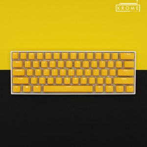 Peut inclure: Un clavier blanc avec des touches jaunes. Le clavier a une disposition standard avec des touches de fonction, des touches numériques et des touches alphabétiques. Le clavier est sur un fond jaune et noir.