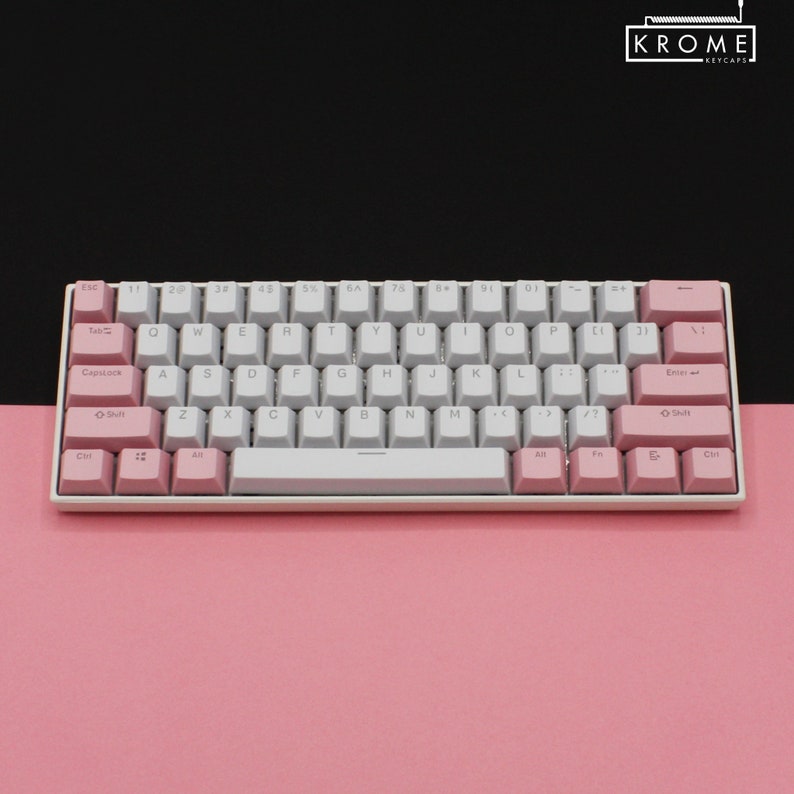May include: A white and pink mechanical keyboard with a white space bar and pink keys. The keyboard has a white frame and is on a pink surface. The keys have white lettering and the top row of keys has black lettering. The keyboard has the word "KROME" in black lettering on the top right corner.