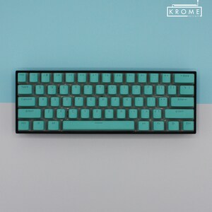 May include: A compact, black keyboard with teal keycaps. The keys are arranged in a standard QWERTY layout. The keyboard has a sleek, minimalist design, with the brand name "KROME KEYCAPS" in the top right corner.
