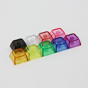 May include: Assortment of ten transparent keycaps in various colours, including black, white, yellow, green, blue, purple, and red. The keycaps are made of a clear plastic material and have a slightly raised edge around the perimeter.