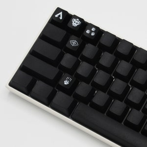 May include: A black mechanical keyboard with white keycaps featuring various symbols and icons, including a skull, a paw print, and an eye. The keyboard has a white frame and a black space bar.
