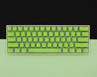 以下が含まれることがあります： 緑色のキーキャップが付いた白いキーボード。キーには白い文字が書かれています。キーボードにはスペースバーとバックスペースキーがあります。キーボードは緑色の表面の上に置かれています。
