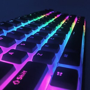 Op de afbeelding: Een wit mechanisch toetsenbord met zwarte toetsen en regenboogkleurige LED-achtergrondverlichting. Het toetsenbord is schuin geplaatst en de toetsen zijn gelabeld met letters en cijfers.