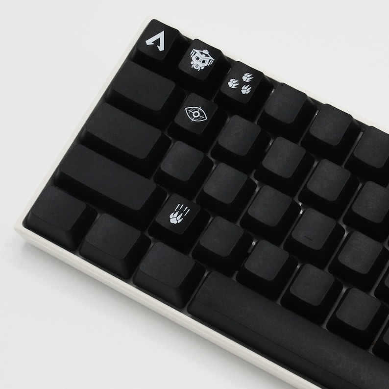 Pode incluir: Um teclado mec&acirc;nico preto com teclas brancas que apresentam v&aacute;rios s&iacute;mbolos e &iacute;cones, incluindo uma caveira, uma pegada de pata e um olho. O teclado tem uma moldura branca e uma barra de espa&ccedil;o preta.