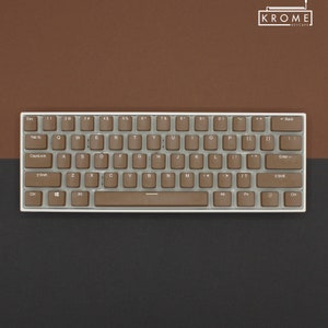 May include: A brown and white mechanical keyboard with the brand name "KROME" printed on the top right corner. The keyboard has a standard layout with keys labeled with letters, numbers, and symbols. The keys are made of a light brown plastic and have a white background.