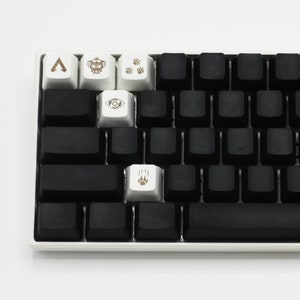 Peut inclure: Un clavier m&eacute;canique noir avec des touches blanches comportant divers symboles, notamment une fl&egrave;che, un robot, des empreintes de pattes et un &oelig;il. Le clavier a une barre d'espace noire.