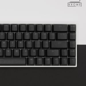 May include: A black mechanical keyboard with white keycaps and a white frame. The keyboard has a standard layout with function keys, arrow keys, and a number pad. The keyboard is on a white surface.