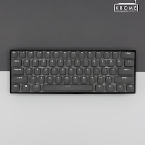 May include: A black and gray mechanical keyboard with a gray keycap set. The keyboard has a standard layout with function keys, arrow keys, and a number pad. The keys are labeled with white lettering. The keyboard is on a gray surface.