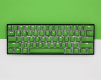 以下が含まれることがあります： 黒枠の緑色のメカニカルキーボード。キーボードは、ファンクションキー、数字キー、矢印キーなど、標準的なレイアウトになっています。キーボードは白い表面の上に置かれています。