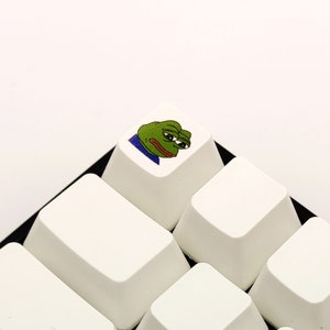 May include: A white keyboard with a single key featuring a green frog meme with a sad expression. The key is surrounded by other blank white keys.