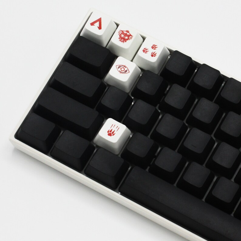 Peut inclure: Un clavier m&eacute;canique blanc avec des touches noires et des touches rouges et blanches avec des symboles. Les symboles comprennent un triangle, un cr&acirc;ne, des empreintes de pattes et un &oelig;il.