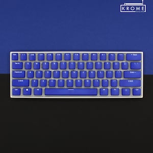 May include: A compact, white-framed mechanical keyboard with blue keycaps. The keys are arranged in a standard QWERTY layout. The background is split between a dark blue and black surface. The upper right corner displays the text "KROME KEYCAPS".