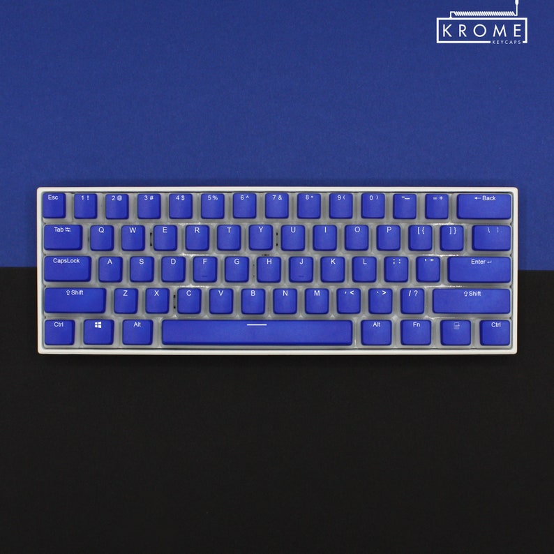 May include: A compact, white-framed mechanical keyboard with blue keycaps. The keys are arranged in a standard QWERTY layout. The background is split between a dark blue and black surface. The upper right corner displays the text "KROME KEYCAPS".