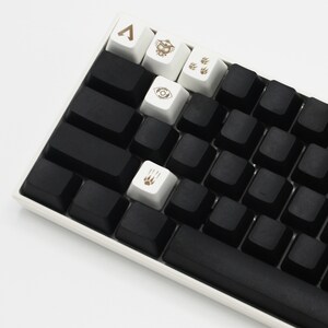 May include: A black mechanical keyboard with white keycaps featuring various symbols, including a triangle, an eye, and paw prints. The keyboard has a black space bar and a white frame.