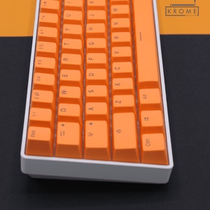 Könnte beinhalten: Eine weiße und orangefarbene mechanische Tastatur mit dem Text "KROME KEYCAPE" in der oberen rechten Ecke.