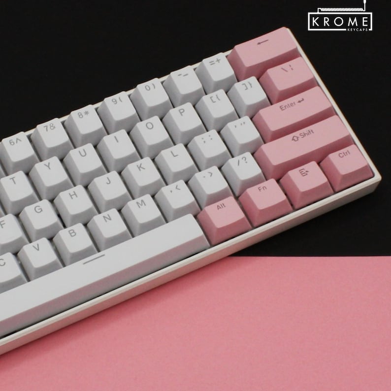 May include: A white mechanical keyboard with pink keycaps. The keyboard has a white space bar and a white frame. The keycaps have the standard layout with letters, numbers, and symbols. The keyboard is on a pink surface.