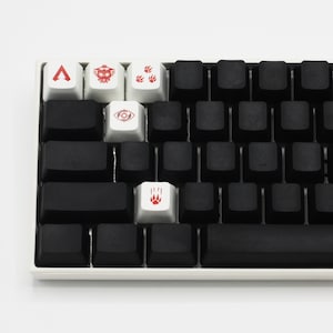 Peut inclure: Un clavier m&eacute;canique blanc avec des touches noires et cinq touches personnalis&eacute;es. Les touches ont des symboles rouges, dont un triangle, un vaisseau spatial, des empreintes de pattes, un &oelig;il et une empreinte de patte.