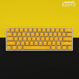 May include: A compact, white-framed keyboard with bright yellow keycaps. The keys are arranged in a standard QWERTY layout. The background is split, with a yellow upper half and a black lower half. The brand name "KROME KEYCAPS" is in the top right corner.