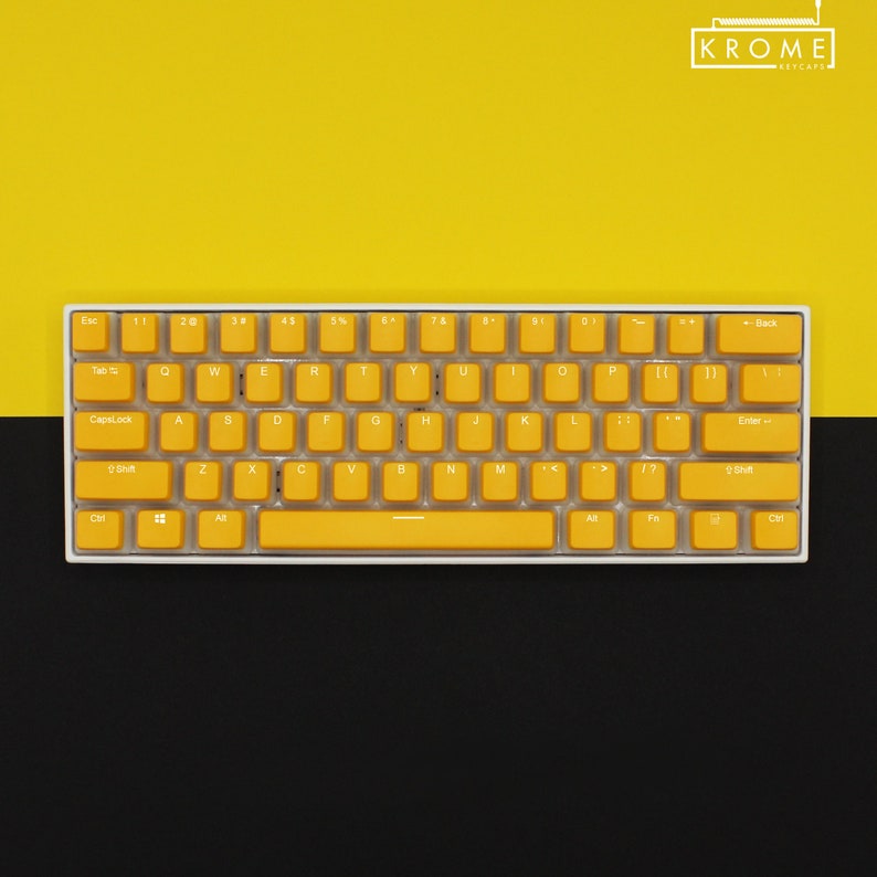 May include: A compact, white-framed keyboard with bright yellow keycaps. The keys are arranged in a standard QWERTY layout. The background is split, with a yellow upper half and a black lower half. The brand name "KROME KEYCAPS" is in the top right corner.