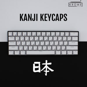 May include: A white mechanical keyboard with Japanese kanji characters on the keys. The keyboard is on a black surface and the word "日本" (Japan) is written in white below the keyboard.