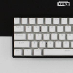 May include: A white mechanical keyboard with black keys and a black frame. The keys have white lettering and symbols. The keyboard has a row of function keys, a row of number keys, a row of letter keys, a row of punctuation keys, and a row of arrow keys. The keyboard also has a space bar, a backspace key, a tab key, a caps lock key, a shift key, a control key, and an alt key.