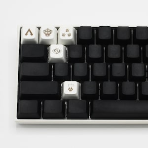 Pode incluir: Um teclado mec&acirc;nico preto com teclas brancas que apresentam v&aacute;rios s&iacute;mbolos, incluindo uma seta, um rob&ocirc;, pegadas de patas e um olho. O teclado tem uma barra de espa&ccedil;o preta.