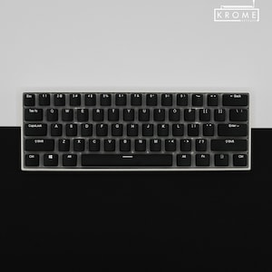 Puede incluir: Un teclado mecánico negro con teclas blancas y un marco blanco. El teclado tiene una disposición estándar con teclas de función, teclas numéricas y una barra espaciadora. El teclado está sobre una superficie negra y blanca.
