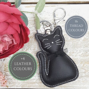 Könnte beinhalten: Schwarzer Leder-Schlüsselanhänger in Katzenform mit weißen Nähten. Der Schlüsselanhänger hat einen silbernen Ring und einen kleinen Anhänger mit der Aufschrift "Handmade with Love". Der Schlüsselanhänger ist von zwei Kreisen umgeben, einer mit dem Text "16 Garnfarben" und der andere mit dem Text "+4 Lederfarben".
