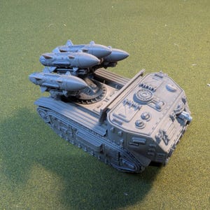 May include: A gray plastic model of a tank with three missiles mounted on a rotating platform. The tank has a detailed design with various hatches and panels.