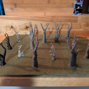 May include: A collection of miniature, stylized trees in various shapes and sizes. Some are black, others are gray, and all are arranged on a green, textured surface. The trees are likely for a model or tabletop game.