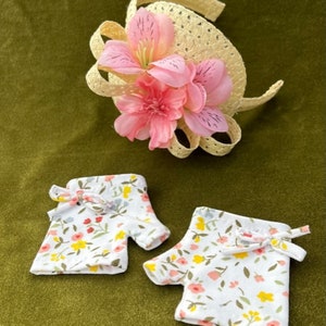 May include: A pair of white fingerless gloves with a floral print in shades of pink, yellow, and green. The gloves have a bow detail at the wrist. A straw headband with a pink flower embellishment is also pictured.