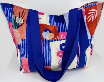 May include: A large tote bag with a colorful patchwork design. The bag features a vibrant mix of patterns, including blue stripes, floral motifs, and abstract shapes in shades of pink, orange, and white. It has sturdy blue straps.