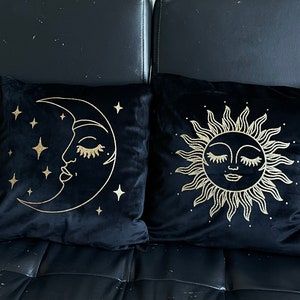 Puede incluir: Dos almohadas de terciopelo negro con bordados dorados. Una almohada presenta una luna creciente con una cara y estrellas. La otra almohada presenta un sol con una cara.
