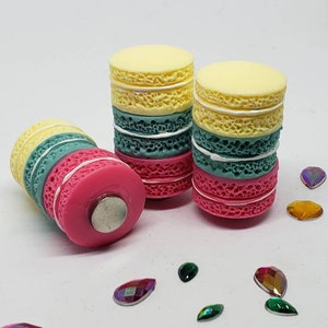 May include: Three miniature macarons stacked on top of each other. The macarons are yellow, green, and pink. The macarons are decorated with white frosting. The macarons are sitting on a white surface with small, iridescent gemstones scattered around them.
