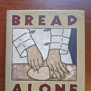 May include: A cookbook titled "BREAD ALONE" with a beige cover. The cover features an illustration of hands kneading dough, with text in red and blue. The book's subtitle reads "BOLD FRESH LOAVES FROM YOUR OWN HANDS".