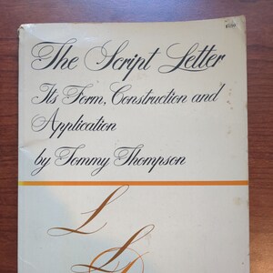 May include: A vintage book titled "The Script Letter: Its Form, Construction and Application" by Tommy Thompson. The cover is off-white with black script lettering and orange accents. Decorative script letter examples are featured.