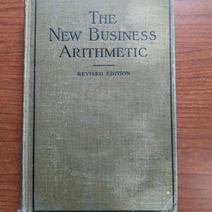 May include: A vintage book titled "The New Business Arithmetic" with a revised edition label. The cover is a muted olive green with black text. The book's cover shows signs of wear, suggesting its age.