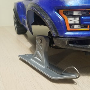 Puede incluir: Un camión de juguete azul con un accesorio de esquí plateado. El camión tiene detalles negros y una textura similar a la tierra. El esquí está unido a la parte delantera del camión y tiene un gancho de metal.