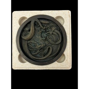 May include: A bronze circular plaque with a raised design of a creature with multiple tentacles and eyes. The plaque is in a white foam packaging.