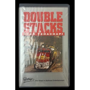 May include: A VHS video cassette with a red and white title card that reads "Double Stacks Over Tehachapi". The cover features a red and yellow train engine with the number 950 on the side. The bottom of the cover has the text "Pentrex... The Finest in Railroad Entertainment".