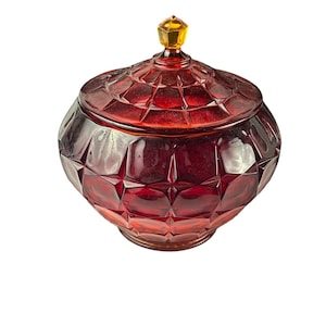 May include: A ruby red, faceted glass jar with a matching lid. The lid is topped with a small, yellow-gold finial. The jar has a round, bulbous shape and is likely used for storage or display. The glass has a diamond-like pattern.