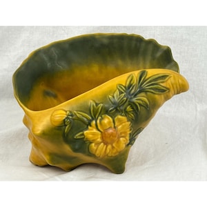 May include: A yellow and green ceramic planter shaped like a seashell with a floral design. The planter features a large yellow flower with green leaves and a smaller yellow flower with green leaves.