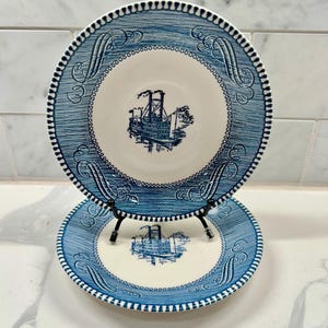 May include: Two round ceramic plates featuring a blue and white transferware pattern. The center of each plate displays a detailed illustration of a paddlewheel steamboat. The wide rim is decorated with blue scrollwork and a repeating scalloped edge.