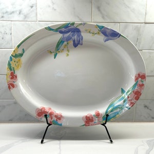 May include: White oval serving platter with a floral design featuring pink, blue, and yellow flowers. The platter is on a black metal stand.