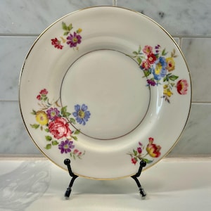 May include: A white porcelain plate with a gold rim and a floral design. The flowers are in shades of red, pink, yellow, blue, and purple. The plate is displayed on a black metal stand.