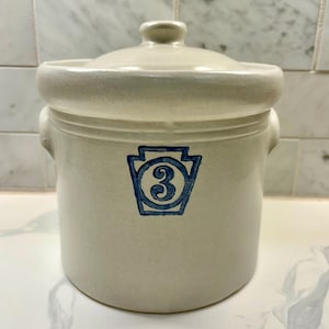 May include: A cream-colored stoneware crock with a matching lid and two side handles. The front features a blue stamped graphic of a keystone shape containing the number 3. The vessel has a smooth, cylindrical body and a thick rim.